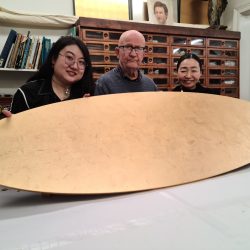 Two students and Karl hold up an oval tabletop which has been gilded with gold leaf.