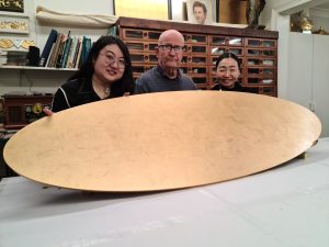 Two students and Karl hold up an oval tabletop which has been gilded with gold leaf.