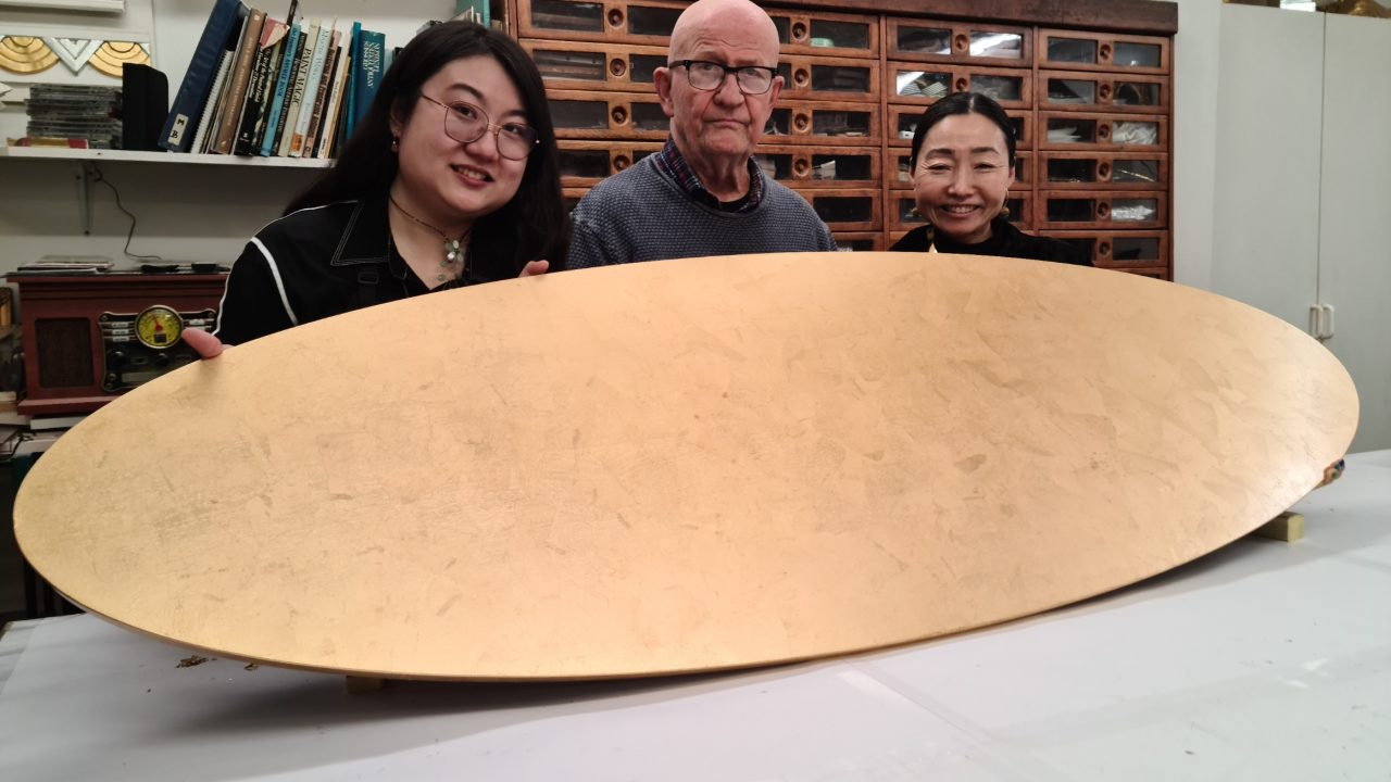 Two students and Karl hold up an oval tabletop which has been gilded with gold leaf.